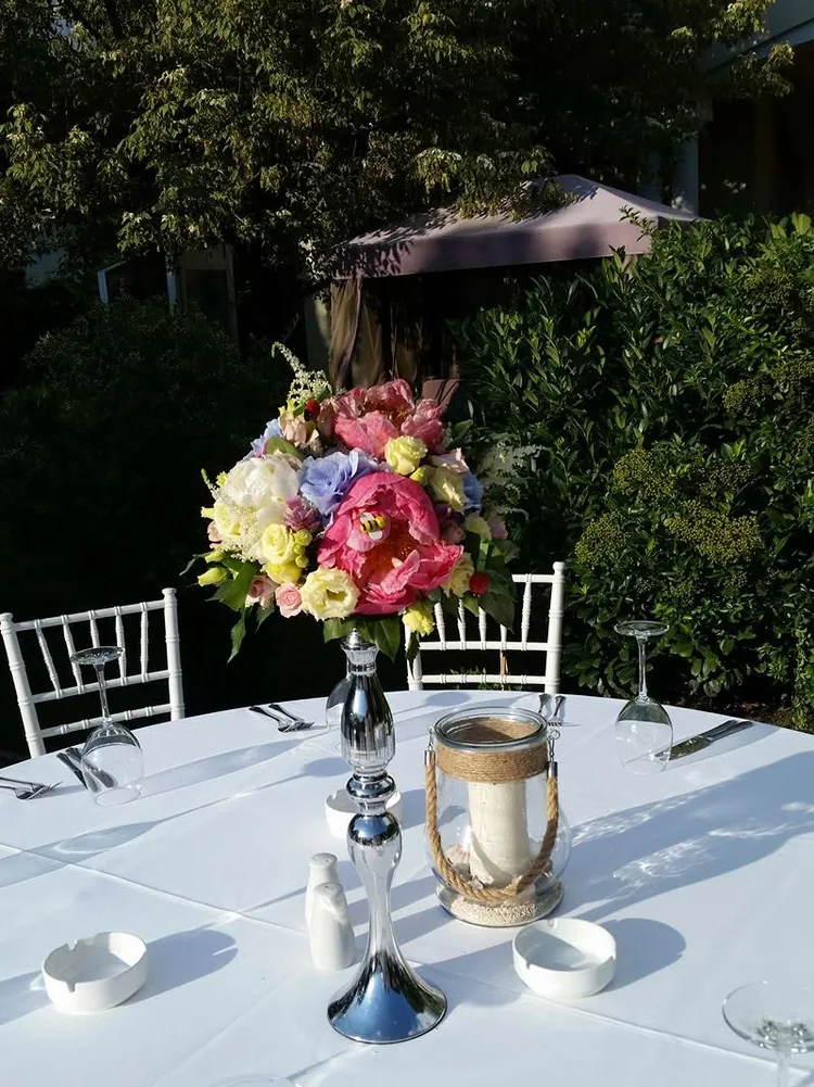 Aranjament înalt pe picior de sticlă, cu bujori roz, hortensie albastră și lisianthus, pe o masă pregătită în grădină